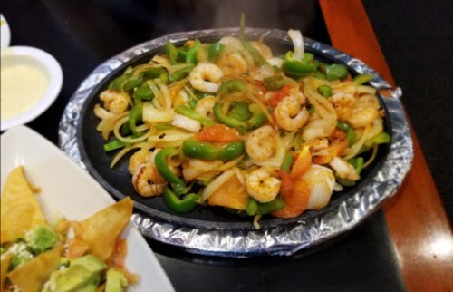 A sizzling platter of shrimp fajitas with bell peppers, onions, and tomatoes, served with sides of chips and guacamole.