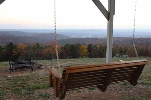 A wooden swing hangs from a structure, overlooking a scenic view of rolling hills and trees at sunset.