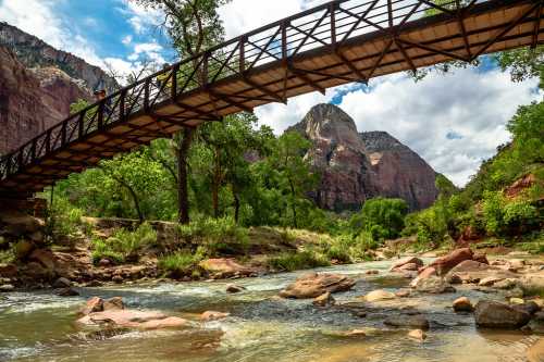 A wooden bridge spans a river surrounded by lush greenery and towering rock formations under a partly cloudy sky.