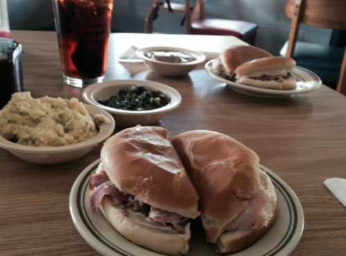 Two sandwiches on plates with sides of mashed potatoes and greens, plus a glass of iced tea on a wooden table.