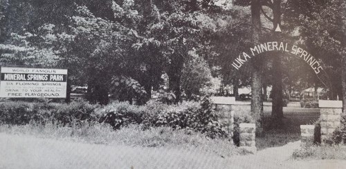 Black and white image of a park entrance sign for Uka Mineral Springs, featuring trees and a stone archway.