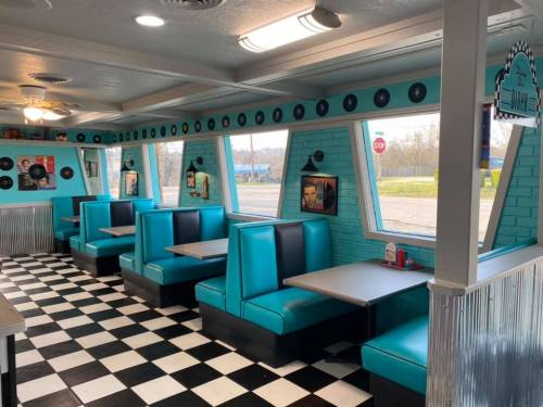 A retro diner interior with teal booths, checkered floor, and large windows, featuring vintage decor and a cozy atmosphere.