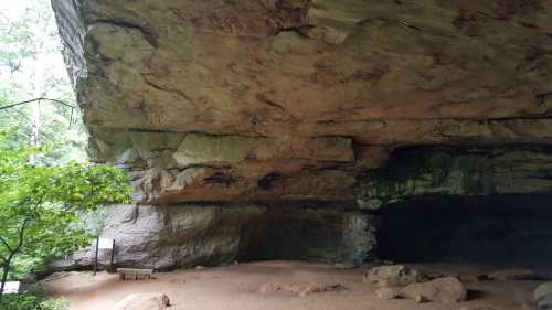 A large rock overhang in a forested area, with sandy ground and a wooden sign in the background.