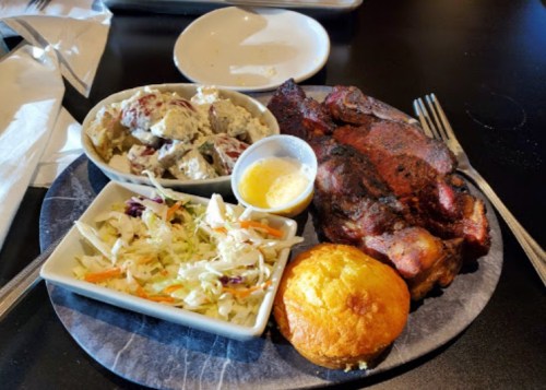 A plate featuring grilled meat, coleslaw, potato salad, a cornbread muffin, and utensils on a dark table.