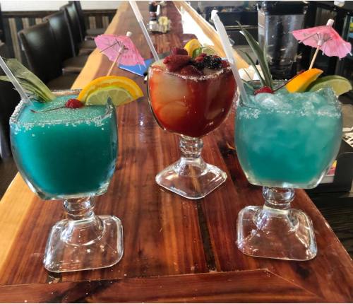 Three colorful cocktails with fruit garnishes and umbrellas, served in decorative glasses on a wooden bar.