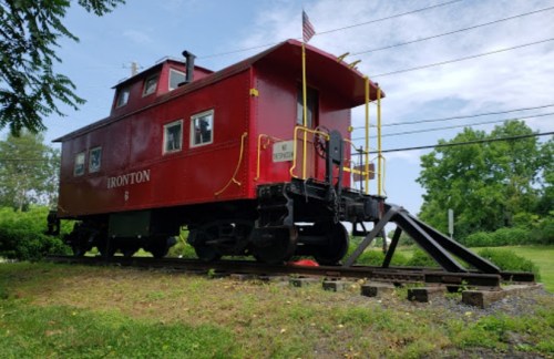 A red caboose labeled "IRONTON" sits on tracks surrounded by greenery under a blue sky.