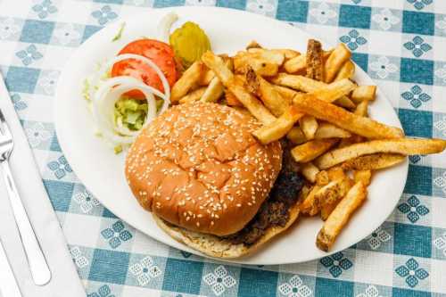 A plate with a sesame seed burger, lettuce, tomato, onion, pickles, and a side of golden French fries on a checkered tablecloth.