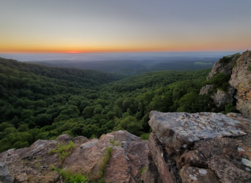A panoramic view of a lush green valley at sunset, with rocky foreground and a colorful sky.