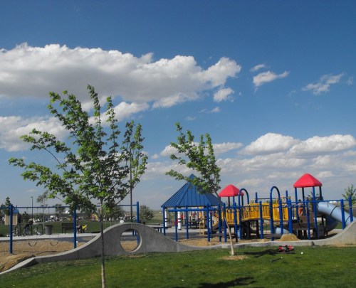 A colorful playground with slides and climbing structures, surrounded by green grass and trees under a blue sky with clouds.