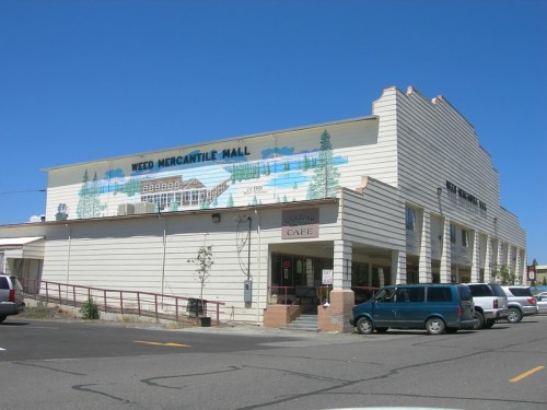 Exterior of Weed Mercantile Mall featuring a mural, with parked cars in front and a clear blue sky.