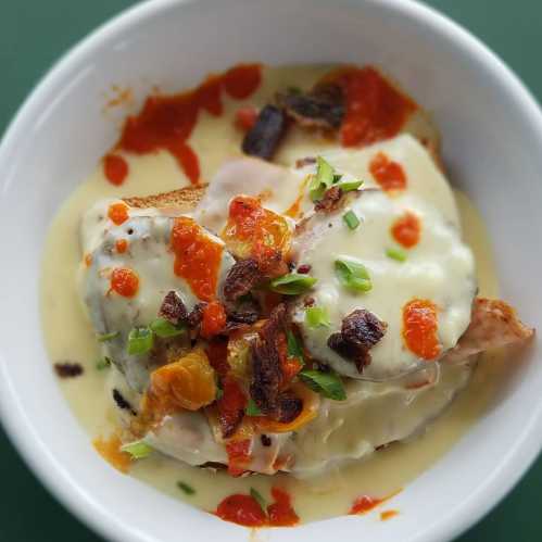 A bowl of creamy sauce topped with bread, melted cheese, chopped green onions, and a drizzle of spicy sauce.