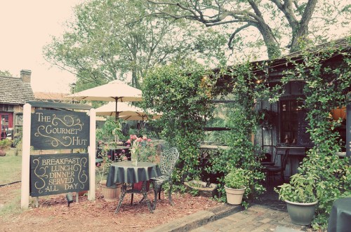 Charming outdoor dining area with a sign for "The Gourmet Hut," surrounded by greenery and tables under umbrellas.