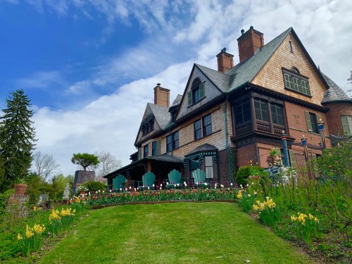 A large, historic house with a garden of colorful flowers under a blue sky, featuring a green lawn and patio chairs.