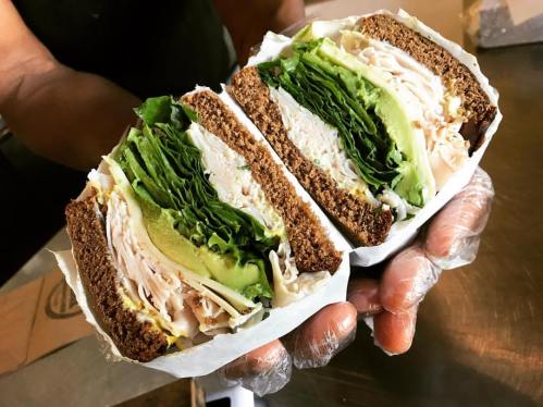 A person holding a sandwich with layers of turkey, lettuce, and avocado on dark bread, wrapped in paper.