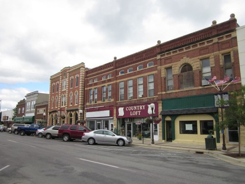 A quaint downtown street featuring brick buildings, shops, and parked cars under a cloudy sky.