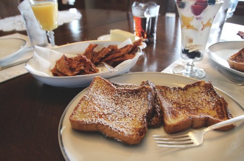 A plate of French toast dusted with powdered sugar, alongside bacon, a parfait, and drinks on a dining table.