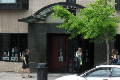 Two women walk near a modern building entrance with large windows and a green tree in front. A car is parked nearby.