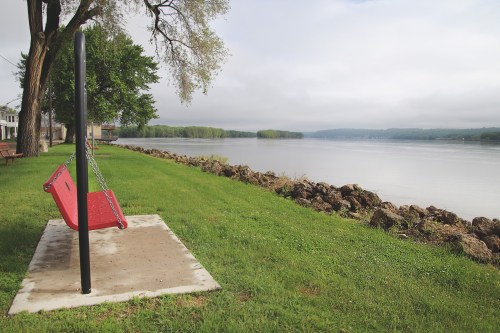 A red swing hangs from a black frame by a calm river, surrounded by green grass and trees under a cloudy sky.