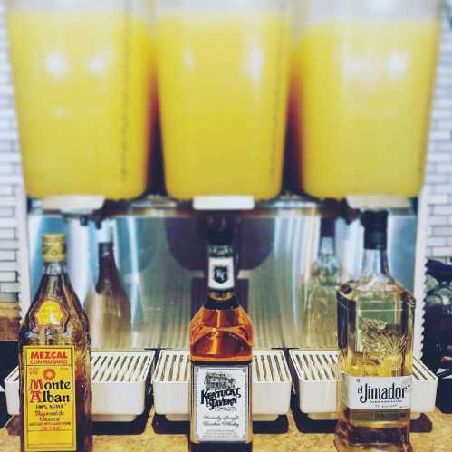 Three bottles of liquor in front of large dispensers filled with yellow liquid, likely cocktails or mixed drinks.