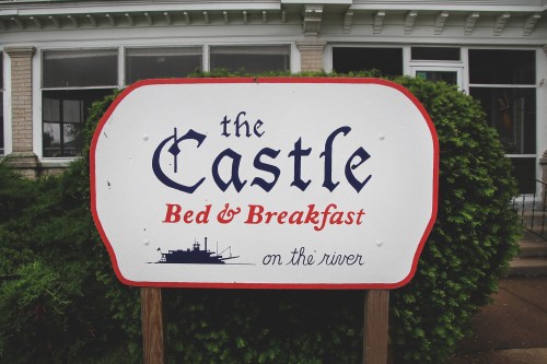 Sign for "The Castle Bed & Breakfast" featuring a boat graphic, located by the river.