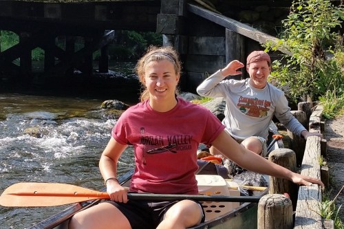Two people in a canoe on a river, smiling and enjoying a sunny day, with greenery and a bridge in the background.
