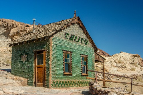 A unique green bottle house with a wooden roof, set against a clear blue sky and rocky landscape.