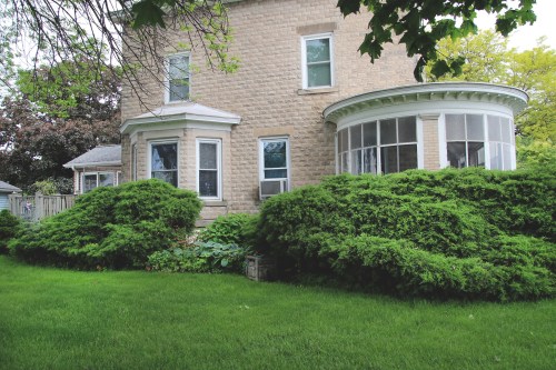 A stone house with a curved porch, surrounded by lush green bushes and a well-maintained lawn.