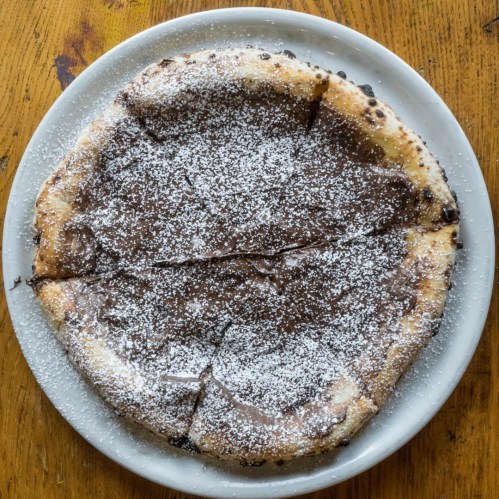 A round dessert pizza topped with chocolate spread and dusted with powdered sugar, sliced into six pieces.