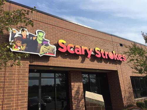 Sign for "Scary Strokes" featuring cartoon monsters, set against a brick building with trees nearby.