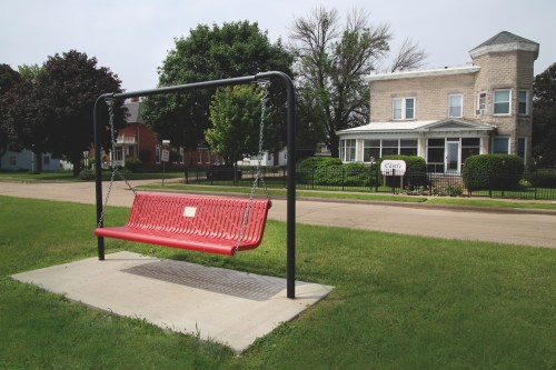 A large red swing on a concrete base in front of a house, with green grass and trees in the background.