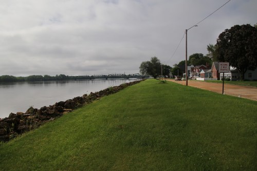A grassy riverbank with a calm water view, lined by trees and a small town in the background under a cloudy sky.