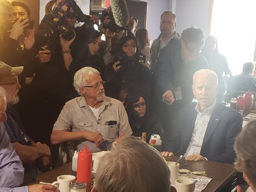 A crowded diner scene with Joe Biden seated at a table, surrounded by people and cameras capturing the moment.