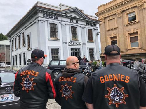 Three bikers in black vests with "Goonies" on the back stand in front of the Oregon Film Museum.