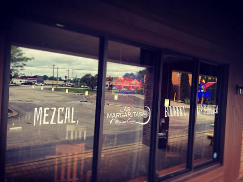 Window view of a restaurant featuring signs for mezcal, margaritas, and bourbon, with an empty parking lot outside.
