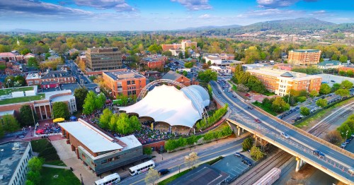Aerial view of a vibrant city with a large outdoor venue, surrounded by greenery and buildings under a blue sky.