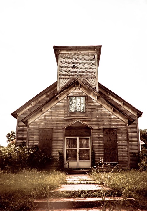 Abandoned wooden church with boarded windows and a weathered sign, surrounded by overgrown grass.