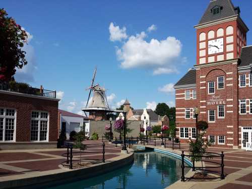 A scenic view of a hotel with a clock tower, a windmill, and a canal, surrounded by flowers and blue skies.