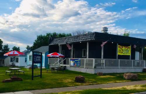 A small restaurant with a black roof, outdoor seating, and flags, featuring a "Now Open" sign on a sunny day.