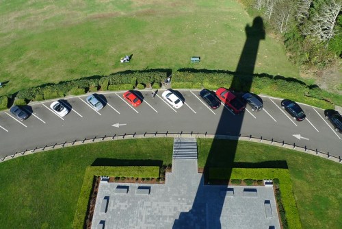 Aerial view of a parking lot with cars and a long shadow cast by a tall structure, surrounded by green grass.