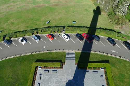 Aerial view of a parking lot with cars and a long shadow cast by a tall structure, surrounded by green grass.