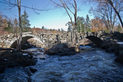 A stone bridge arches over a rushing river, surrounded by trees and autumn foliage under a clear blue sky.