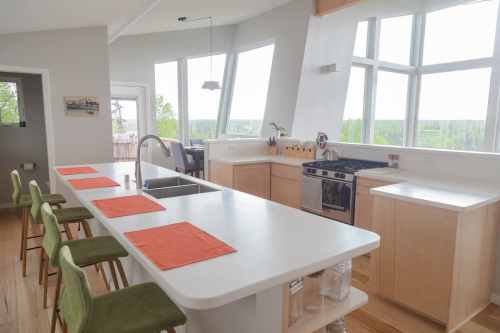 Modern kitchen with large windows, a central island, green bar stools, and a sleek design. Bright and airy atmosphere.
