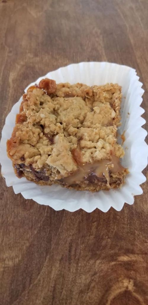A square dessert bar with a crumbly oat topping and a gooey caramel layer, placed in a white paper liner.