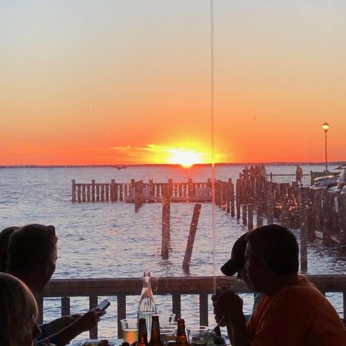 A sunset over a calm waterway, with silhouettes of people dining at a table and a pier in the background.