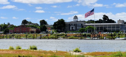 A scenic view of a riverbank with buildings, trees, and an American flag waving in the foreground under a blue sky.