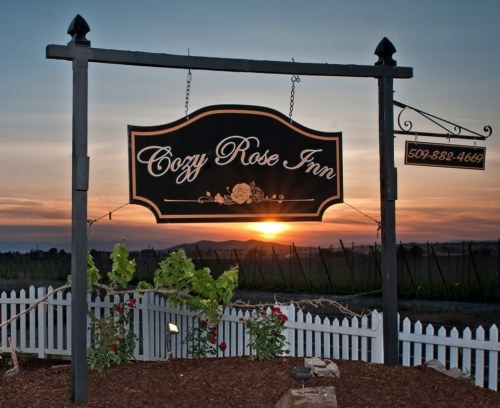 Sign for Cozy Rose Inn at sunset, surrounded by flowers and a white picket fence, with mountains in the background.