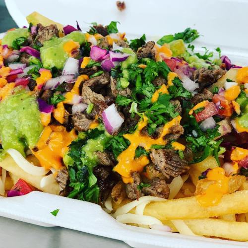 A close-up of loaded fries topped with steak, cheese, onions, cilantro, and green sauce in a takeout container.