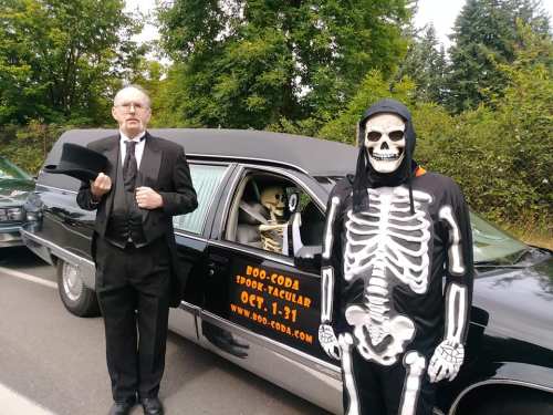 A man in a suit and a skeleton costume stand by a hearse with a skeleton inside, promoting a Halloween event.