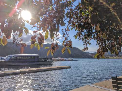 A serene lakeside view with boats, mountains in the background, and sunlight filtering through leafy branches.