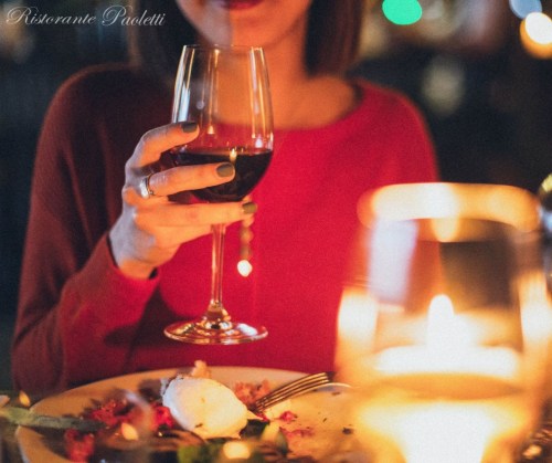 A woman in a red sweater holds a glass of red wine, with a candlelit dinner setting in the background.